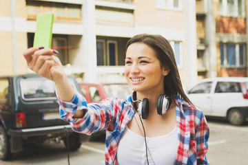 Young woman taking a selfie outdoors in urban area. Teenage girl vlogging outside, smiling, wearing plaid shirt. Female person with smartphone and headphones