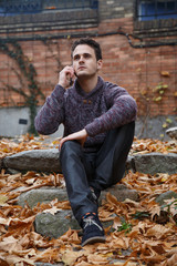 Young handsome man sitting on a rock in meadow with foliage and speaking with smartphone 