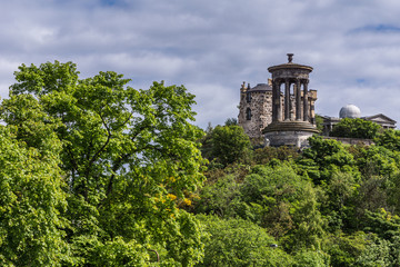 Edinburgh, Scotland, UK - June 13, 2012: Seen from Old Calton Cemetery. Donald Stewart memorial peeks above green tree foliage under blue cloudy sky. 