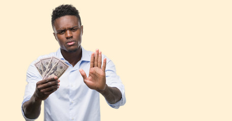 Young african american businessman holding dollars with open hand doing stop sign with serious and...