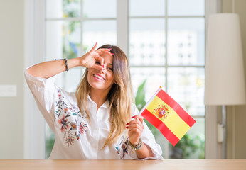 Young woman at home holding flag of Spain with happy face smiling doing ok sign with hand on eye looking through fingers