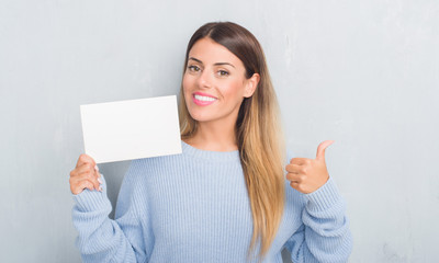 Naklejka premium Young adult woman over grey grunge wall holding blank card pointing and showing with thumb up to the side with happy face smiling