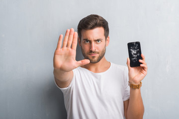 Handsome young man over grey grunge wall showing broken smartphone screen with open hand doing stop sign with serious and confident expression, defense gesture