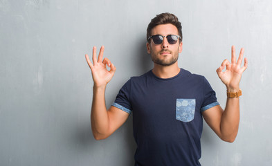 Naklejka premium Handsome young man over grey grunge wall wearing sunglasses relax and smiling with eyes closed doing meditation gesture with fingers. Yoga concept.