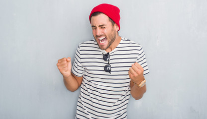 Naklejka premium Handsome young hipster man over grey grunge wall wearing navy t-shirt and wool cap very happy and excited doing winner gesture with arms raised, smiling and screaming for success. Celebration concept.