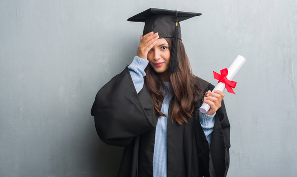 Young brunette woman over grunge grey wall wearing graduate uniform holding degree stressed with hand on head, shocked with shame and surprise face, angry and frustrated. Fear and upset for mistake.