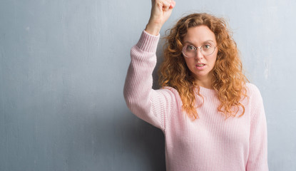 Young redhead woman over grey grunge wall wearing glasses annoyed and frustrated shouting with...