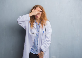 Young redhead doctor woman over grey grunge wall wearing a coat peeking in shock covering face and eyes with hand, looking through fingers with embarrassed expression.