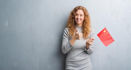 Young redhead woman over grey grunge wall holding flag of China happy with big smile doing ok sign, thumb up with fingers, excellent sign