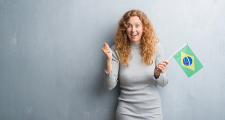 Young redhead woman over grey grunge wall holding flag of Brazil screaming proud and celebrating victory and success very excited, cheering emotion