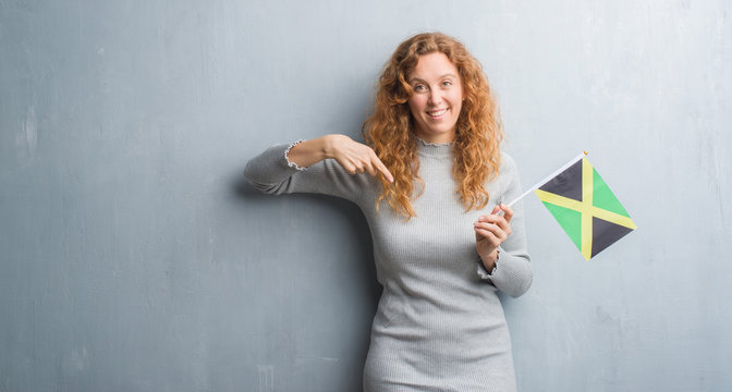 Young redhead woman over grey grunge wall holding flag of Jamaica with surprise face pointing finger to himself
