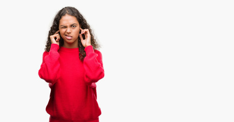 Young hispanic woman wearing red sweater covering ears with fingers with annoyed expression for the noise of loud music. Deaf concept.