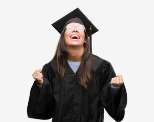 Young hispanic woman wearing graduated cap and uniform very happy and excited doing winner gesture with arms raised, smiling and screaming for success. Celebration concept.