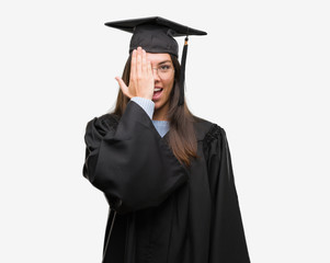 Young hispanic woman wearing graduated cap and uniform covering one eye with hand with confident smile on face and surprise emotion.