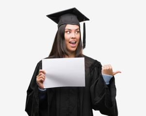 Young hispanic woman wearing graduated uniform holding diploma paper pointing and showing with thumb up to the side with happy face smiling