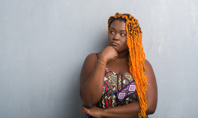 Young african american woman over grey grunge wall wearing orange braids with hand on chin thinking about question, pensive expression. Smiling with thoughtful face. Doubt concept.