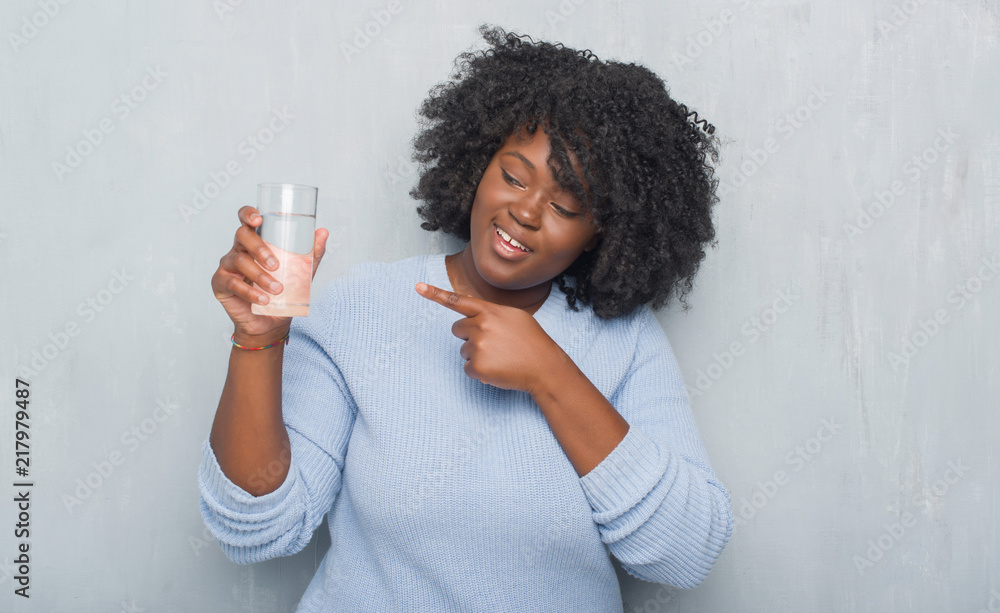 Poster Young african american woman over grey grunge wall drinking a glass of water very happy pointing with hand and finger