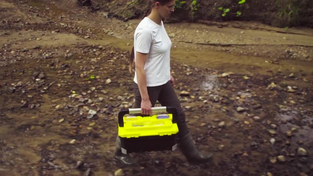 Dolly shot. The woman scientist environmentalist with the tool box in her hand going to the place of research near creek. Side view.