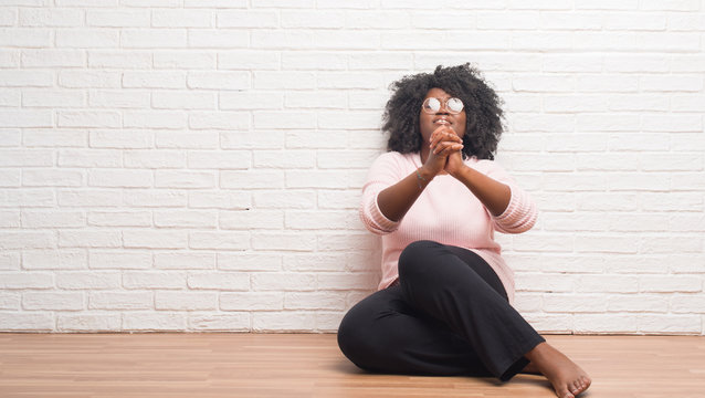 Young african american woman sitting on the floor at home begging and praying with hands together with hope expression on face very emotional and worried. Asking for forgiveness. Religion concept.