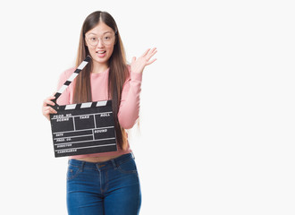 Young Chinese woman over isolated background holding clapper very happy and excited, winner expression celebrating victory screaming with big smile and raised hands