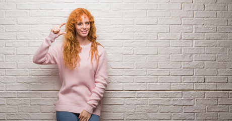 Young redhead woman standing over brick wall smiling and confident gesturing with hand doing size sign with fingers while looking and the camera. Measure concept.