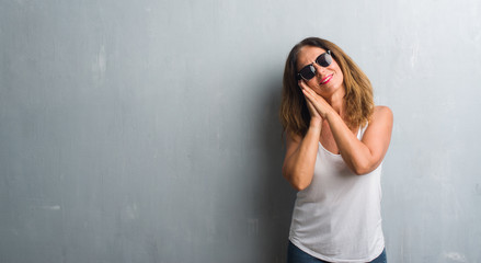 Middle age hispanic woman over grey wall wearing sunglasses sleeping tired dreaming and posing with hands together while smiling with closed eyes.