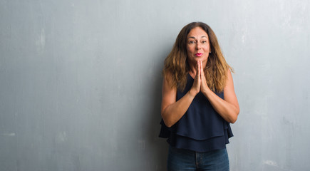 Fototapeta premium Middle age hispanic woman standing over grey grunge wall praying with hands together asking for forgiveness smiling confident.
