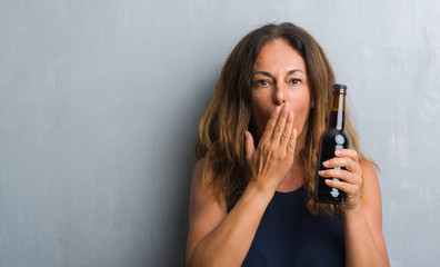 Fototapeta premium Middle age hispanic woman standing over grey grunge wall holding beer bottle cover mouth with hand shocked with shame for mistake, expression of fear, scared in silence, secret concept