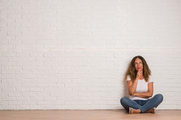 Middle age hispanic woman over white brick wall serious face thinking about question, very confused idea