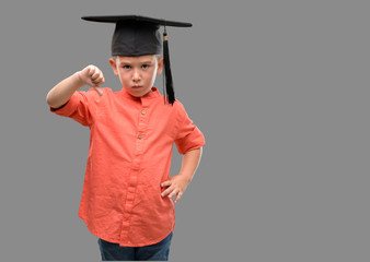 Dark haired little child wearing graduation cap with angry face, negative sign showing dislike with thumbs down, rejection concept