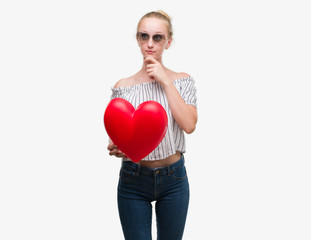 Blonde teenager woman holding red heart serious face thinking about question, very confused idea