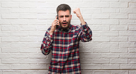 Young adult man talking on the phone standing over white brick wall annoyed and frustrated shouting with anger, crazy and yelling with raised hand, anger concept