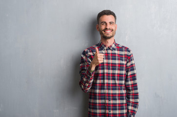 Young adult man standing over grey grunge wall happy with big smile doing ok sign, thumb up with fingers, excellent sign