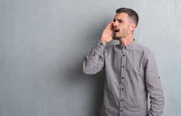 Young adult man standing over grey grunge wall shouting and screaming loud to side with hand on mouth. Communication concept.