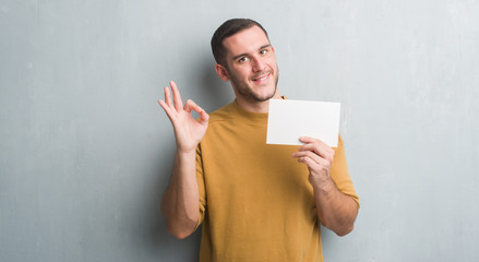 Young caucasian man over grey grunge wall holding blank card doing ok sign with fingers, excellent symbol