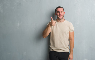 Young caucasian man over grey grunge wall doing happy thumbs up gesture with hand. Approving expression looking at the camera with showing success.