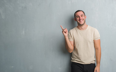 Young caucasian man over grey grunge wall with a big smile on face, pointing with hand and finger to the side looking at the camera.