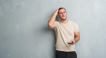 Young caucasian man over grey grunge wall texting a message using smartphone stressed with hand on head, shocked with shame and surprise face, angry and frustrated. Fear and upset for mistake.