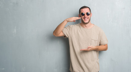 Young caucasian man over grey grunge wall wearing sunglasses gesturing with hands showing big and large size sign, measure symbol. Smiling looking at the camera. Measuring concept.