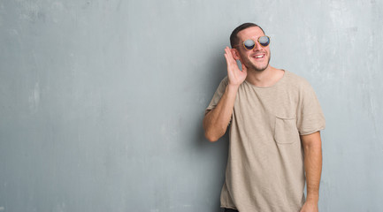 Young caucasian man over grey grunge wall wearing sunglasses smiling with hand over ear listening an hearing to rumor or gossip. Deafness concept.