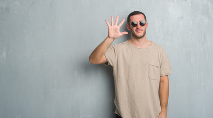 Young caucasian man over grey grunge wall wearing sunglasses showing and pointing up with fingers number five while smiling confident and happy.