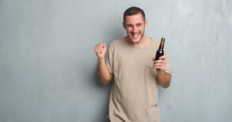 Young caucasian man over grey grunge wall holding bottle beer screaming proud and celebrating victory and success very excited, cheering emotion