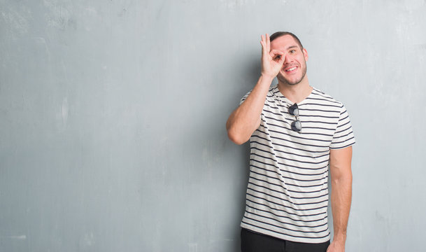 Young caucasian man over grey grunge wall doing ok gesture with hand smiling, eye looking through fingers with happy face.