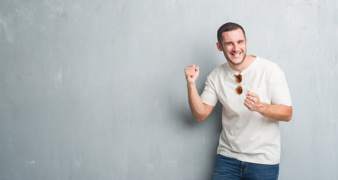 Young caucasian man over grey grunge wall wearing sunglasses very happy and excited doing winner gesture with arms raised, smiling and screaming for success. Celebration concept.