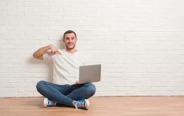 Young caucasian man sitting over white brick wall using computer laptop with surprise face pointing finger to himself