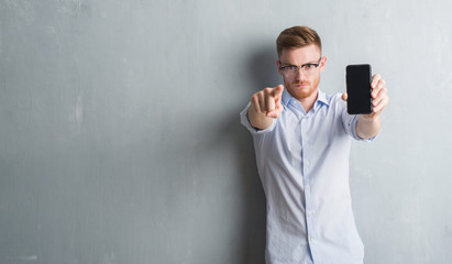 Young redhead man over grey grunge wall showing blank screen of smartphone pointing with finger to the camera and to you, hand sign, positive and confident gesture from the front