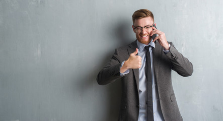 Young redhead  business man over grey grunge wall talking on the phone happy with big smile doing ok sign, thumb up with fingers, excellent sign