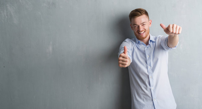 Young redhead business man over grey grunge wall approving doing positive gesture with hand, thumbs up smiling and happy for success. Looking at the camera, winner gesture.