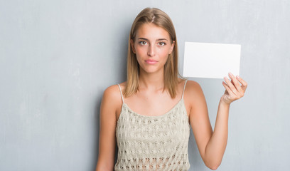 Beautiful young woman over grunge grey wall holding blank card with a confident expression on smart face thinking serious