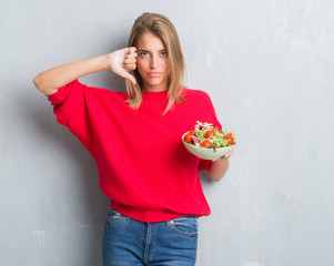 Beautiful young woman over grunge grey wall eating tomato salad with angry face, negative sign showing dislike with thumbs down, rejection concept